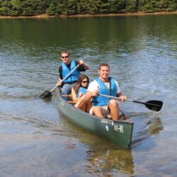 Three smiling people paddle a canoe on a calm lake.