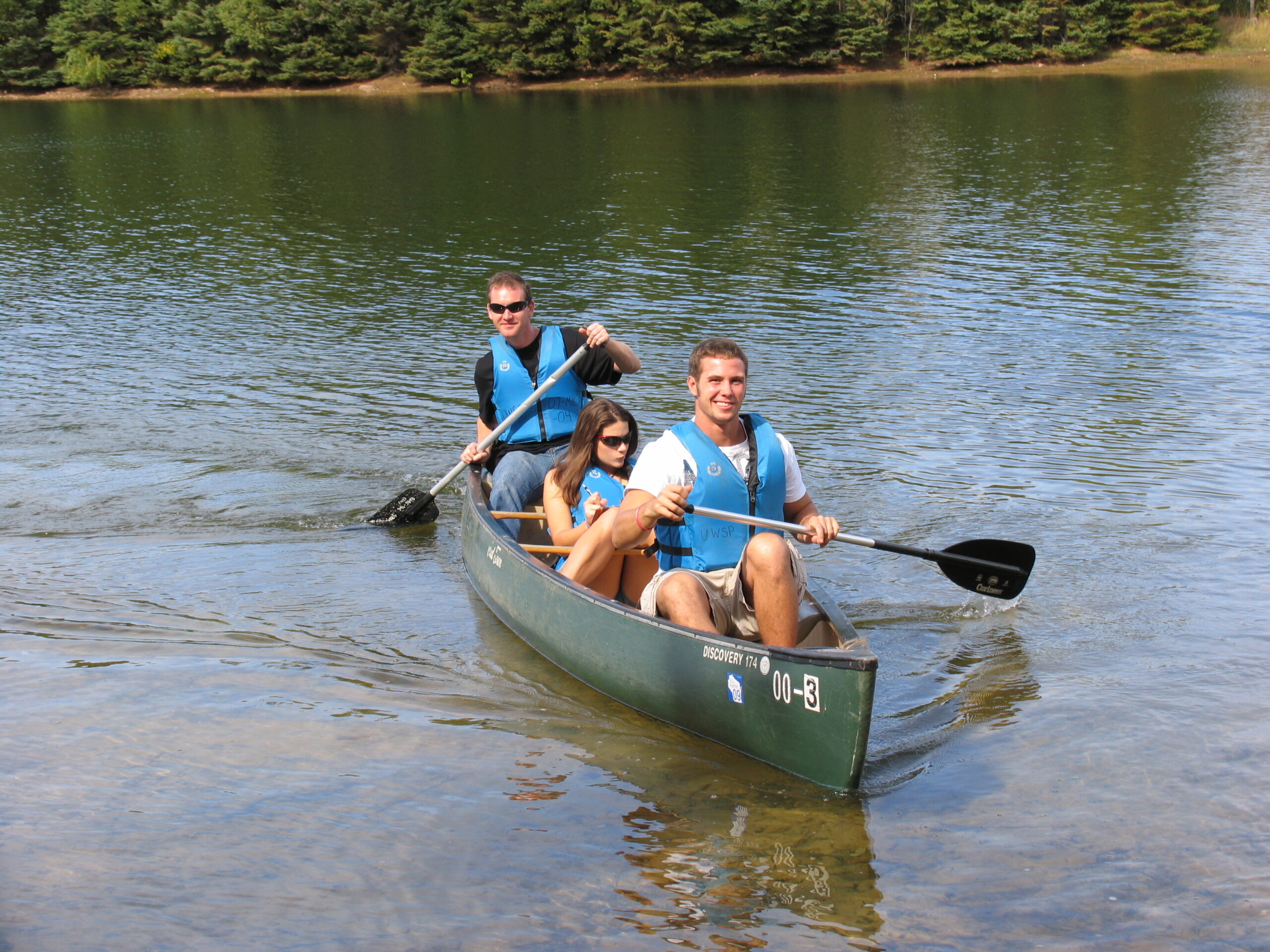 Three smiling people paddle a canoe on a calm lake.