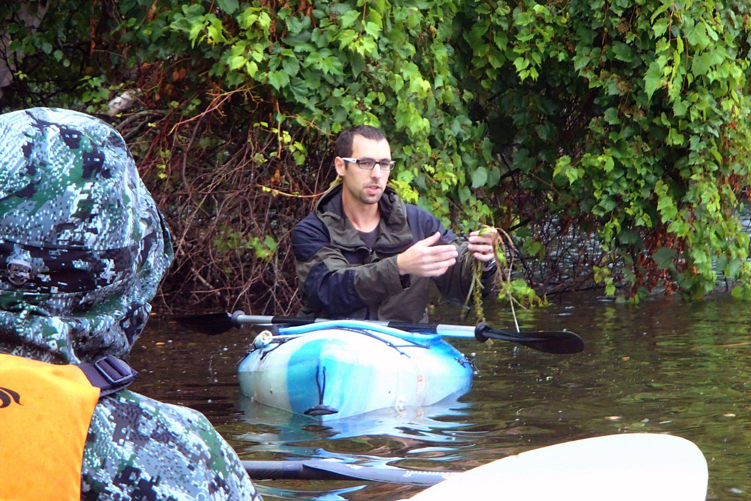 A man in a blue kayak shows an aquatic plant to a group of visitors.