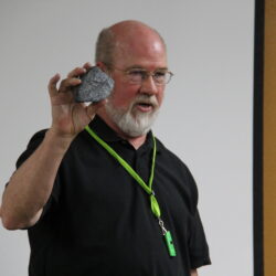 A man holds up a large grey rock in his right hand.